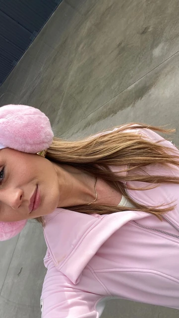 Portrait of a young woman wearing earmuffs and a pink jacket, posing indoors against a gray wall, capturing a modern and fashionable look.