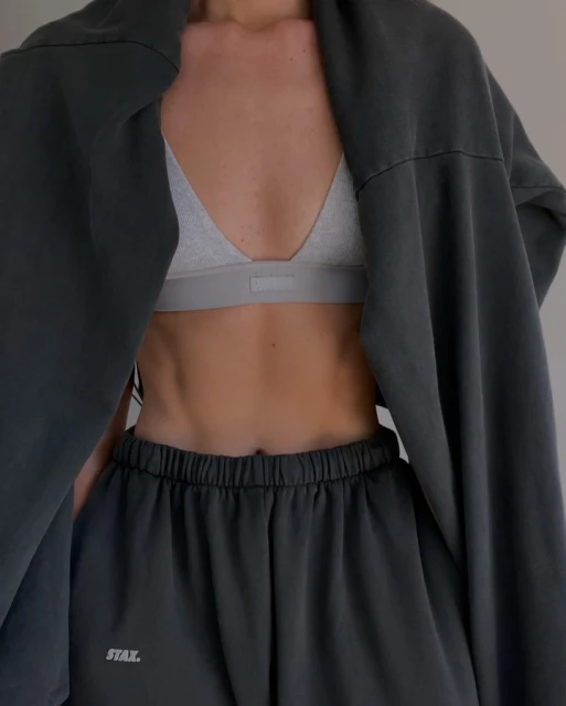 Close-up of a woman wearing a grey sports bra and sweatpants with a sweatshirt draped open over her shoulders in a neutral studio.
