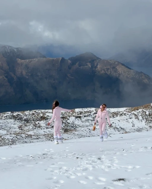 Two adult females in pink ski suits pose in a snowy mountain environment with cloudy skies and distant peaks in the background.
