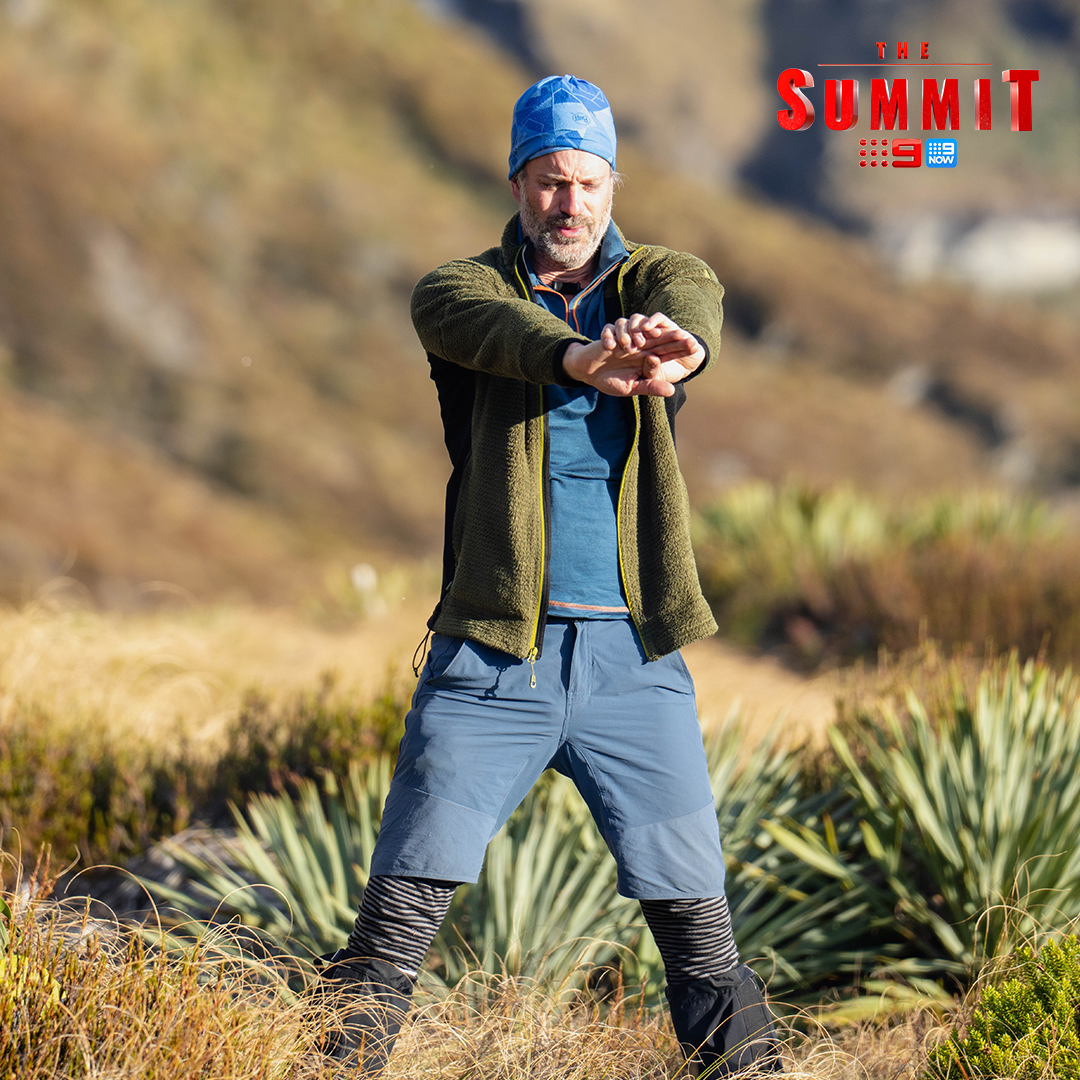 Adult male in athletic wear standing on grassy hillside with mountains in background and text overlay of ‘The Summit’ TV show logo.