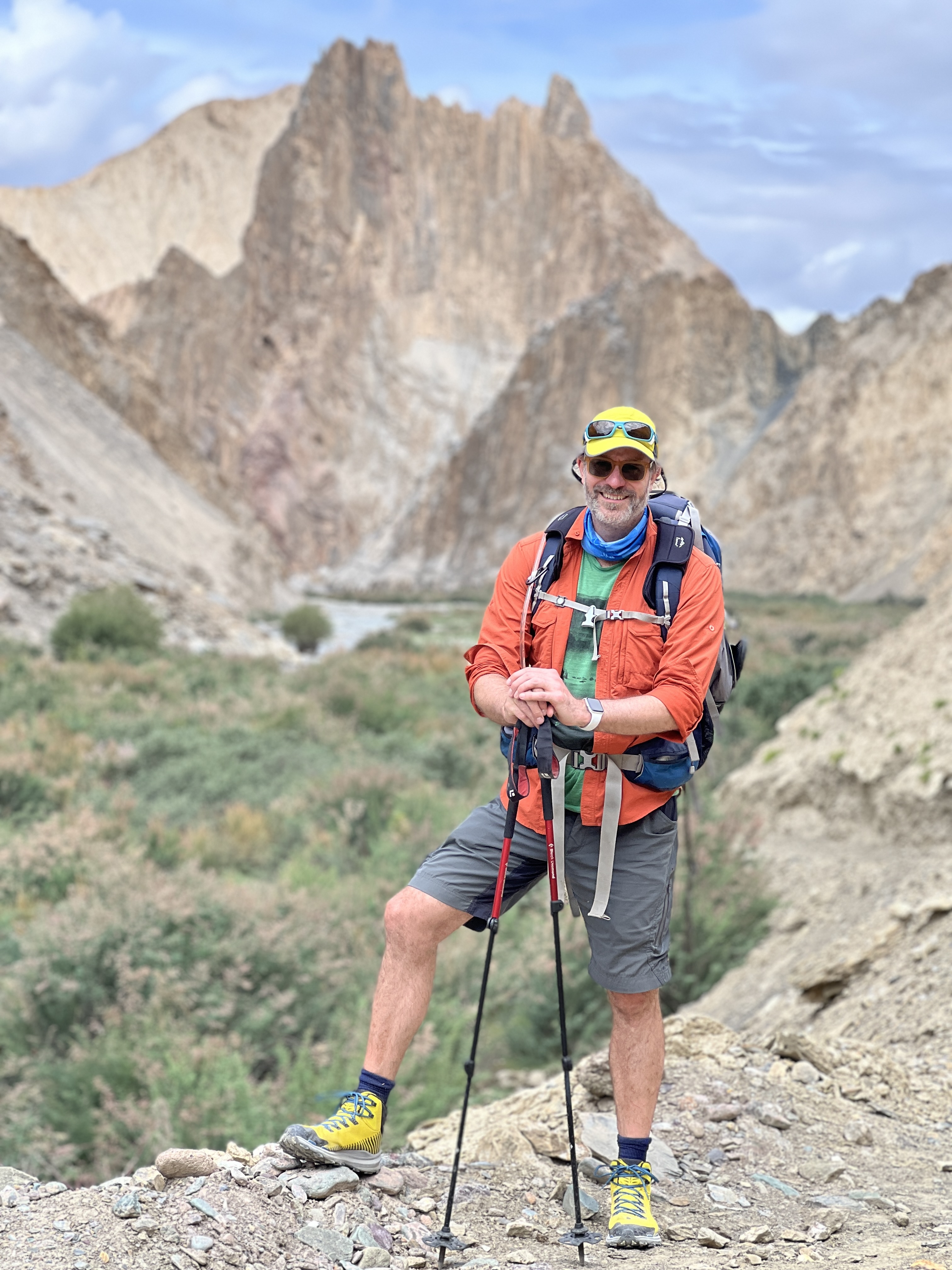 A man hikes in a mountainous landscape wearing a backpack, orange jacket, sunglasses, and hat while using hiking poles for support.