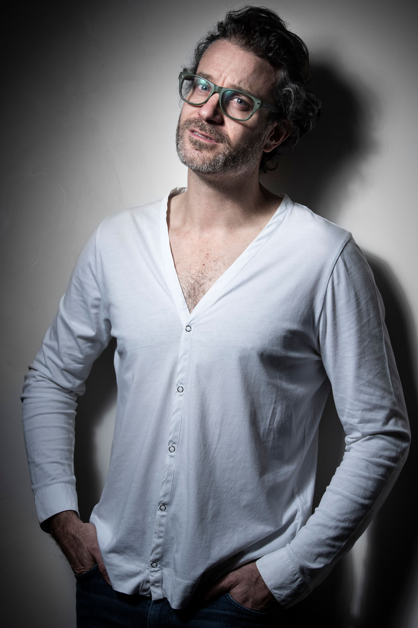 Indoor portrait of a middle-aged Caucasian man with glasses, wearing a white cardigan, hands in pockets, against a neutral gray background.