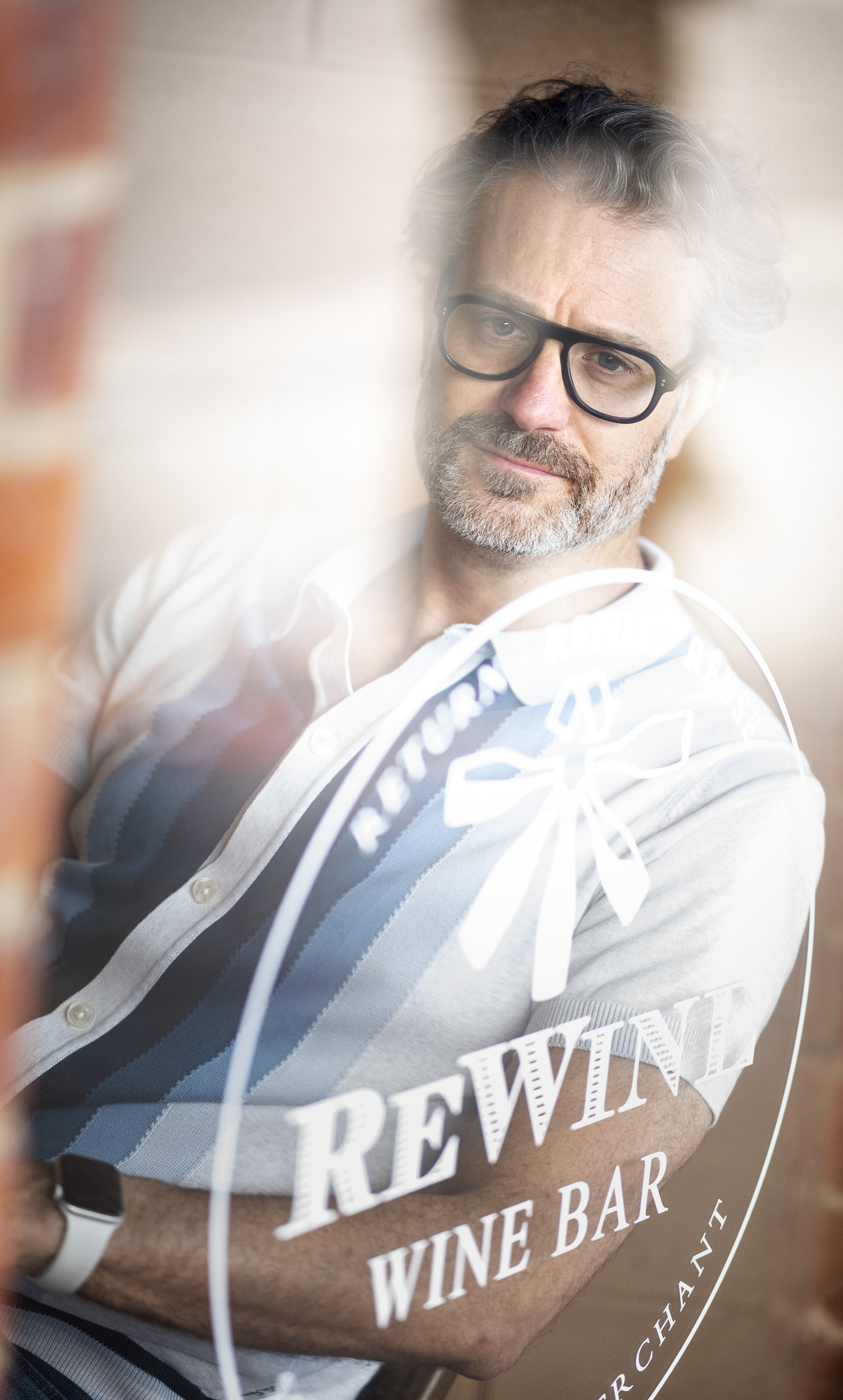 Portrait of a man with glasses in front of a window with the words REWIND WINE BAR on it and a brick wall in the background.
