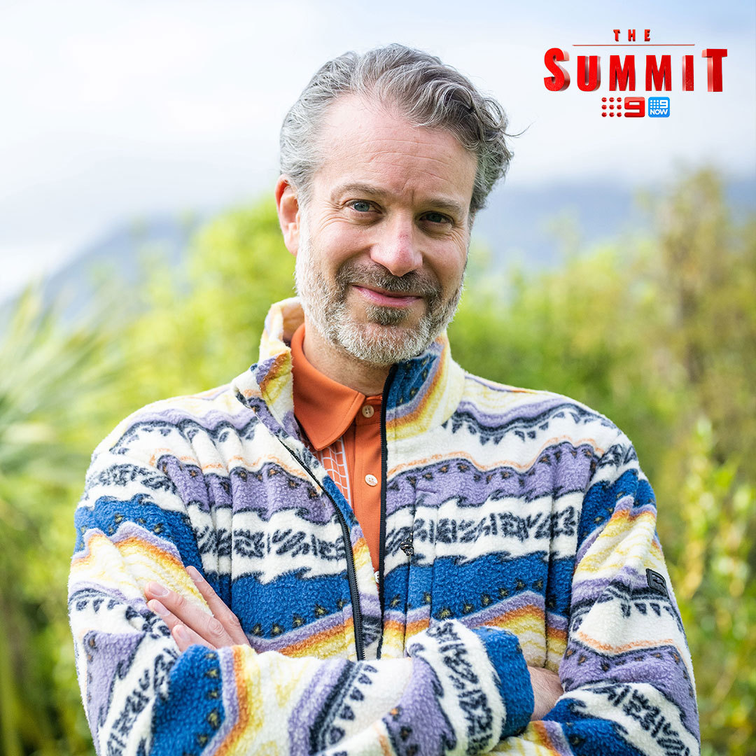Portrait of a smiling adult male in a colorful fleece jacket, arms crossed, with greenery and a mountain visible in the background.