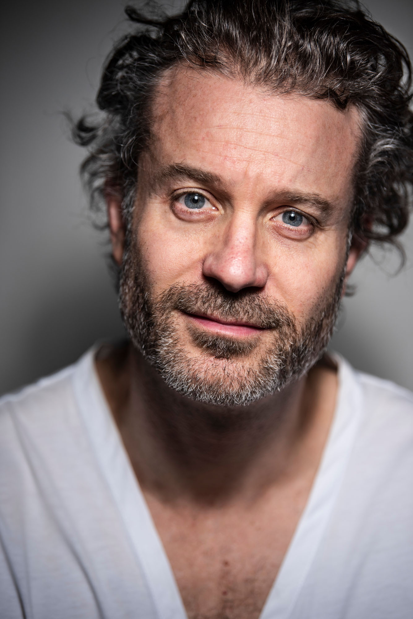 Close-up portrait of a handsome man with curly dark hair, a salt-and-pepper beard, and a white v-neck shirt against a gray background.