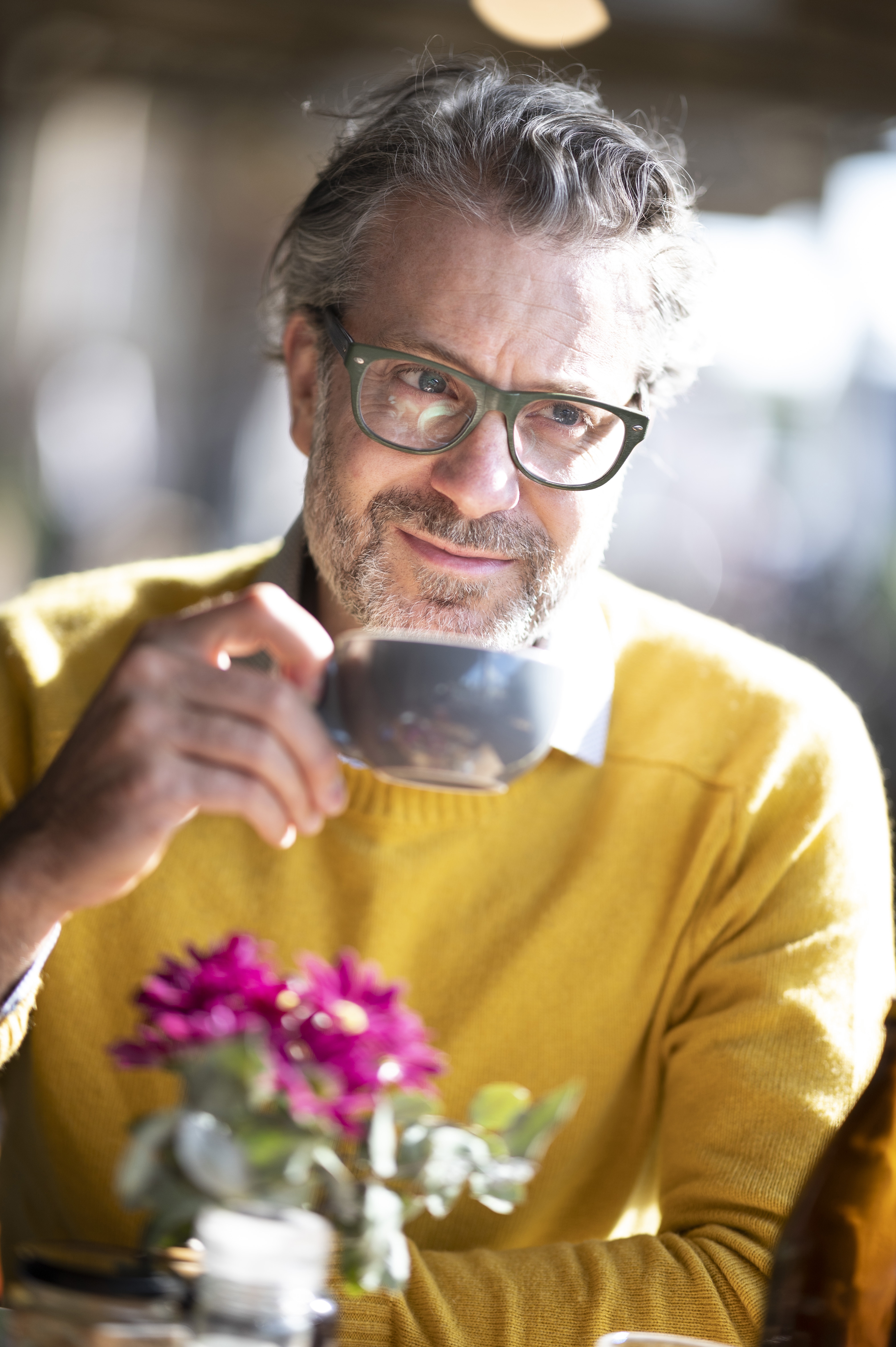 An adult male wearing glasses and a yellow sweater sits at a table with a cup, with flowers, and natural lighting.