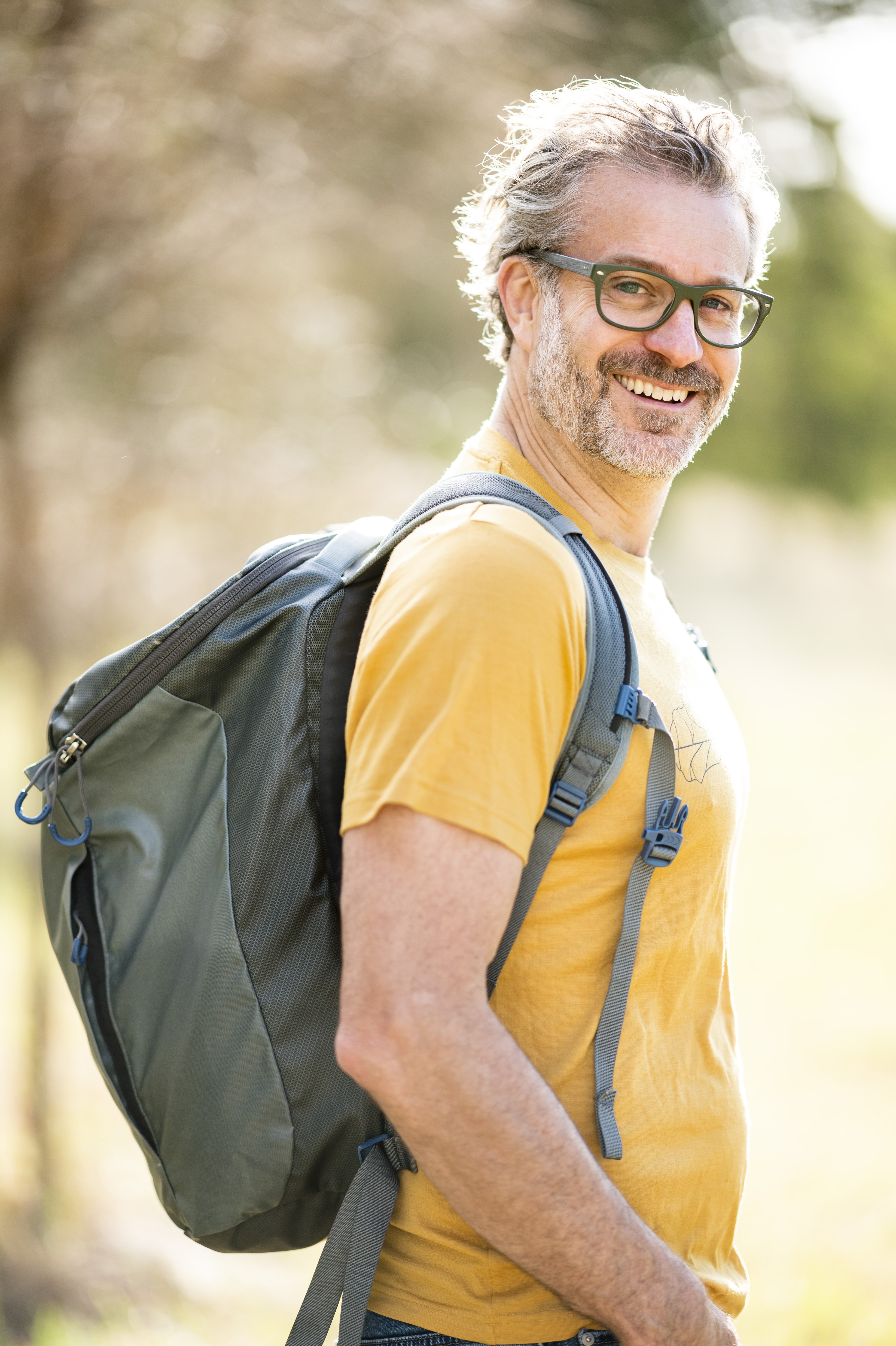 Smiling man with grey hair and glasses wearing a yellow shirt and a backpack in an outdoor setting with blurred background.