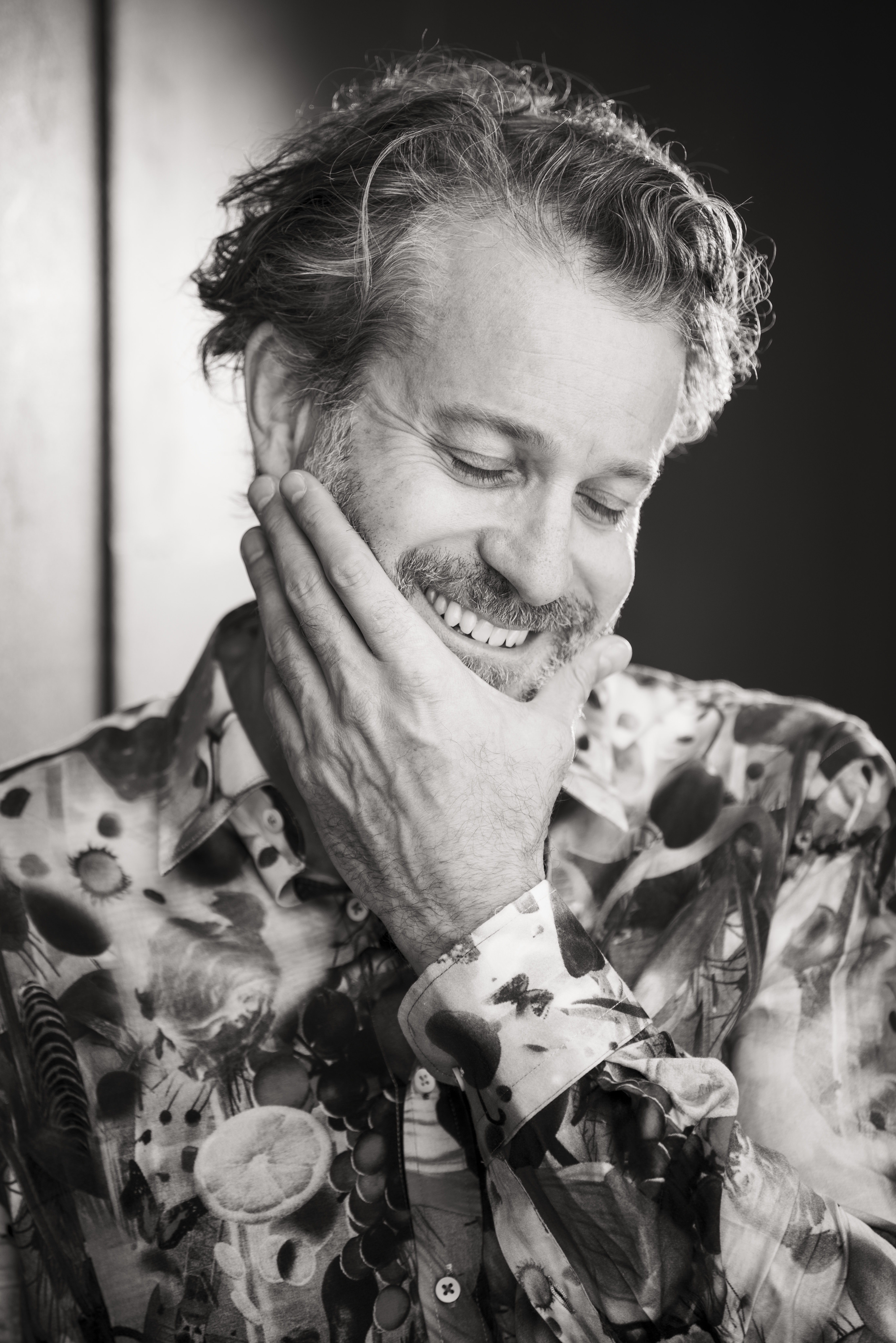 Black and white portrait of a smiling adult male with patterned shirt, hand to face, and a blurred dark background.
