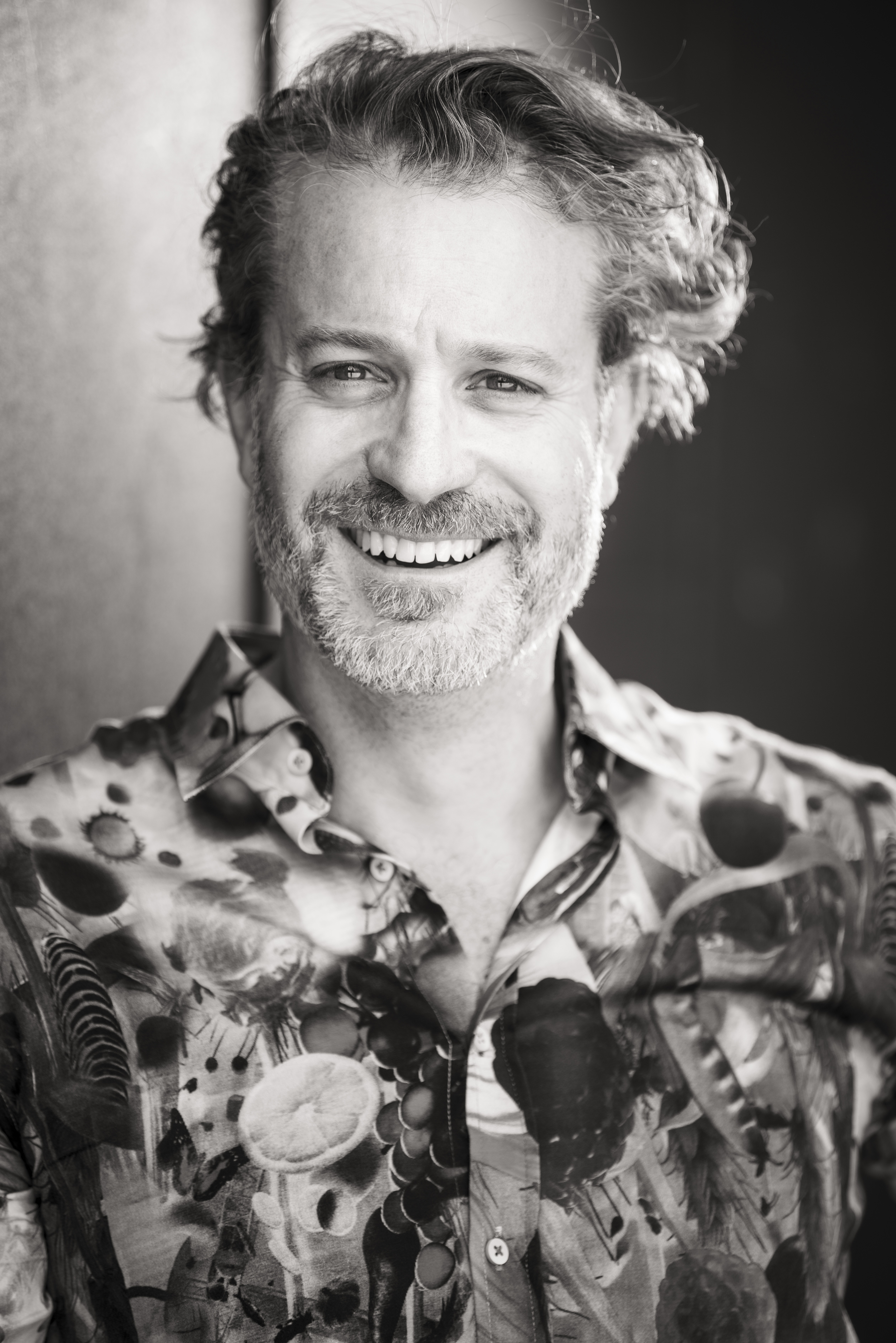 Black and white portrait of an adult male with a beard, smiling, wearing a patterned shirt against a dark backdrop, head and shoulders.