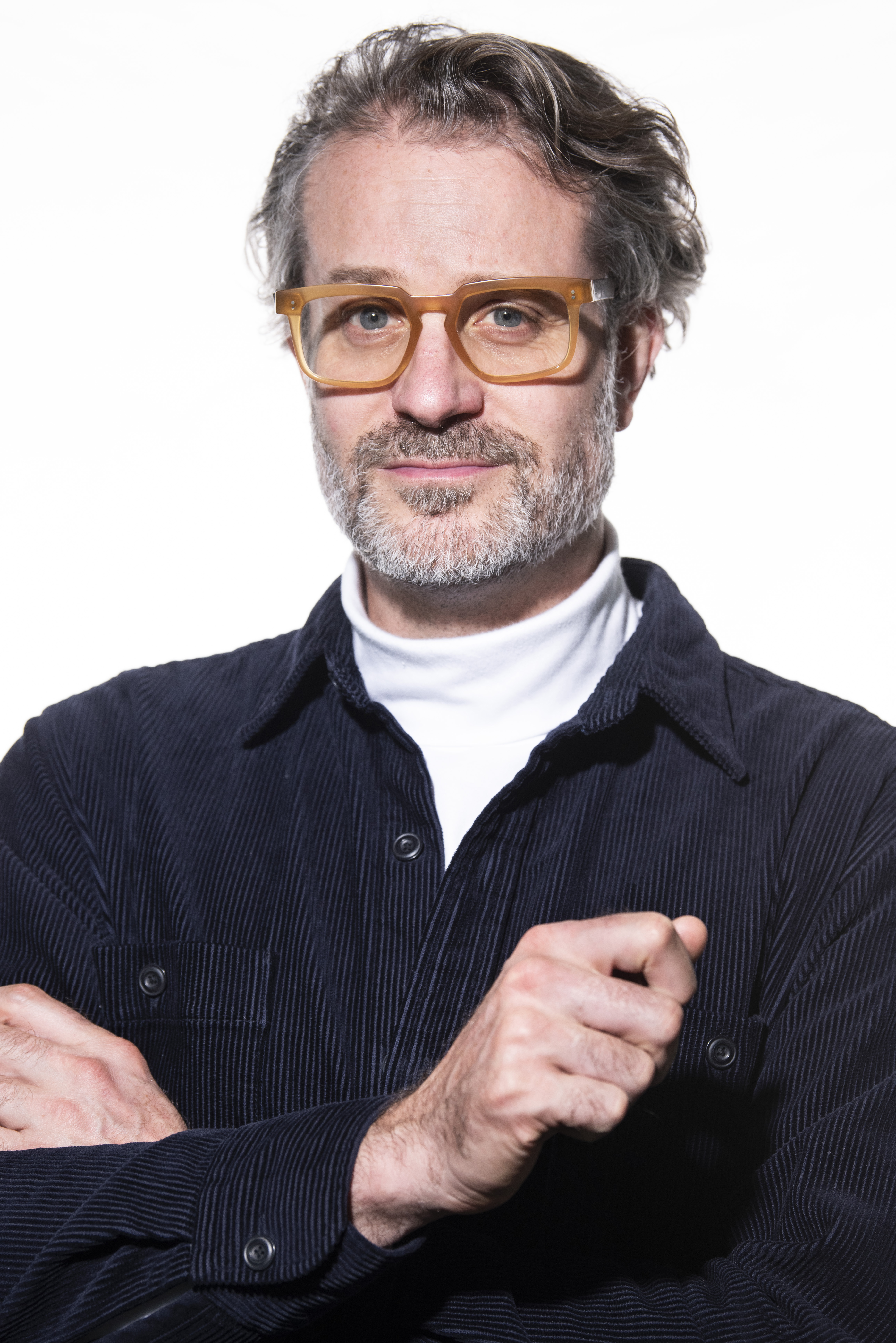 Portrait of a grey-haired man with a beard wearing brown-framed glasses, a white turtleneck and a blue corduroy jacket on a white background.