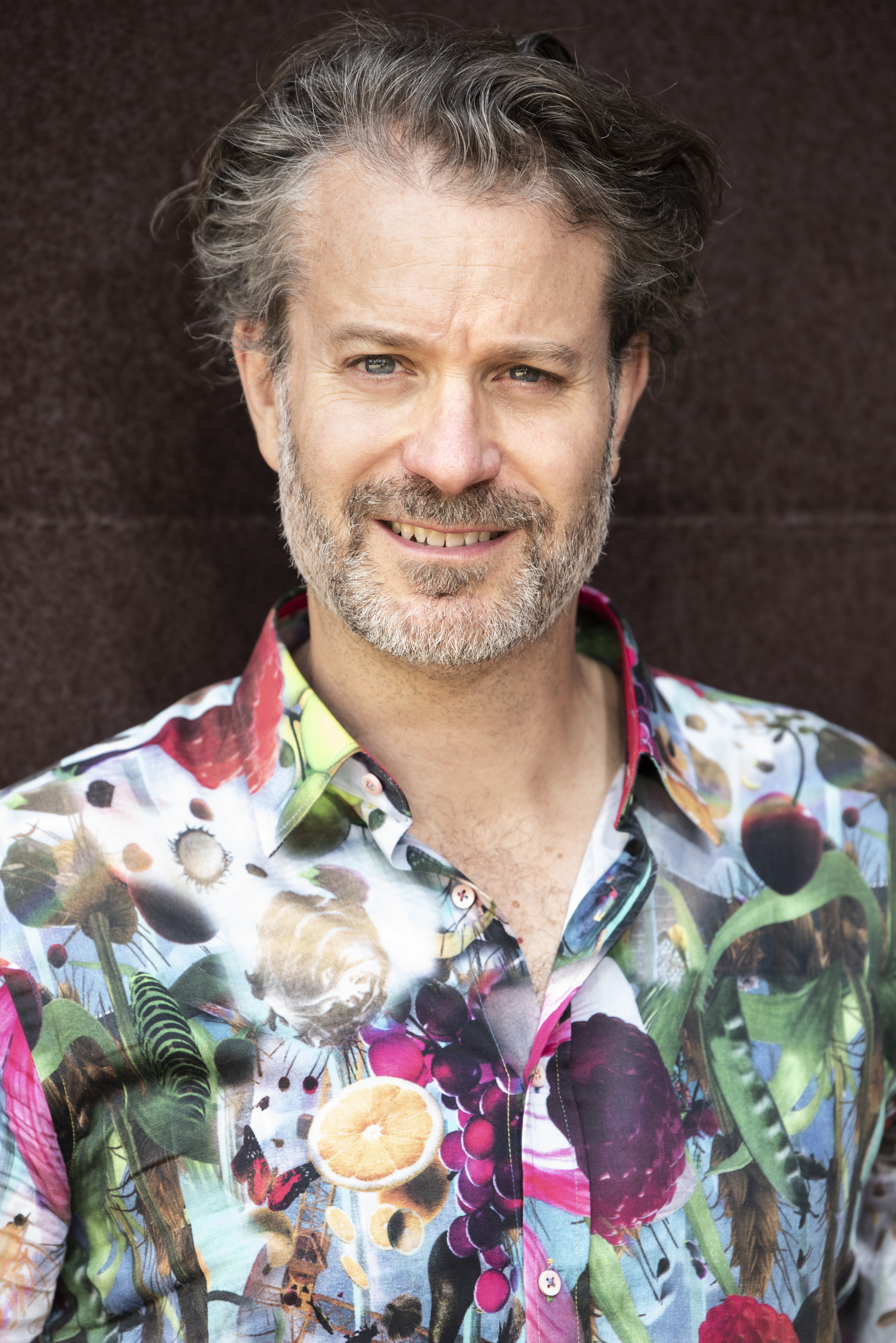 Portrait of an adult male smiling at the camera, wearing a colourful patterned shirt, standing in front of a mottled brown background.
