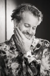 Portrait of a smiling man with wavy hair and beard wearing a patterned button-down shirt, hand on chin