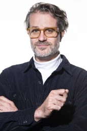 Portrait of a man with glasses wearing a white turtleneck and dark corduroy shirt against a white background