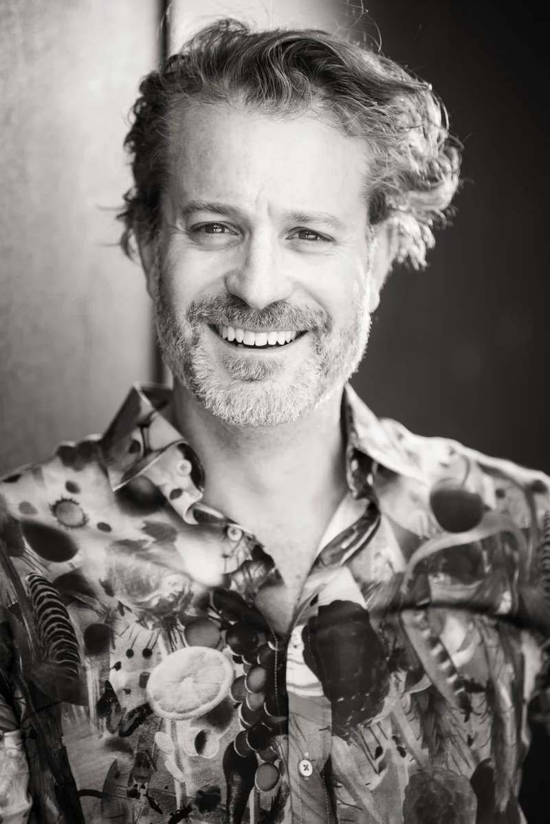 Portrait of a smiling man with curly hair and beard wearing a patterned shirt in a black and white photograph by Josh Waldhorn