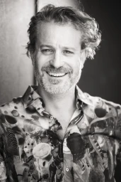 Portrait of a smiling man with curly hair and beard wearing a patterned shirt in a black and white photograph by Josh Waldhorn