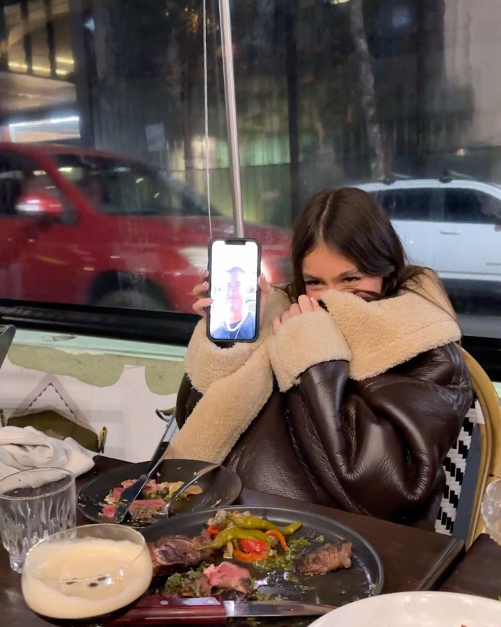 A woman sits at a restaurant table holding up a phone displaying a picture of a person, with steak plates and a car visible through the window.