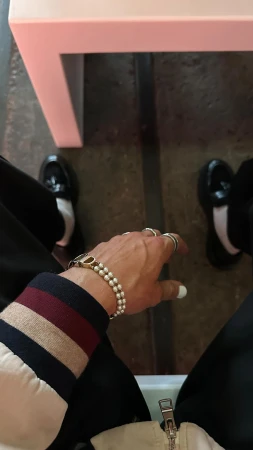 First-person angle of a hand with pearl bracelet and rings, wearing a striped jacket with black trousers and loafers