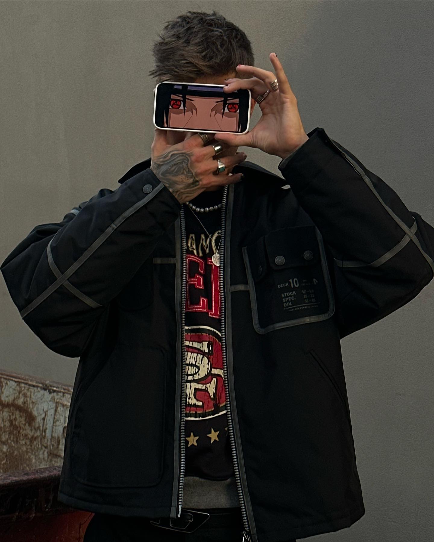 Adult man holding smartphone with anime character eyes on screen. Man wears jacket, t-shirt, rings, and a necklace. Gray backdrop.