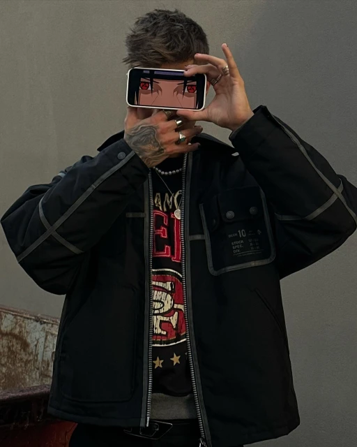 Adult man holding smartphone with anime character eyes on screen. Man wears jacket, t-shirt, rings, and a necklace. Gray backdrop.