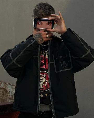 Adult man holding smartphone with anime character eyes on screen. Man wears jacket, t-shirt, rings, and a necklace. Gray backdrop.