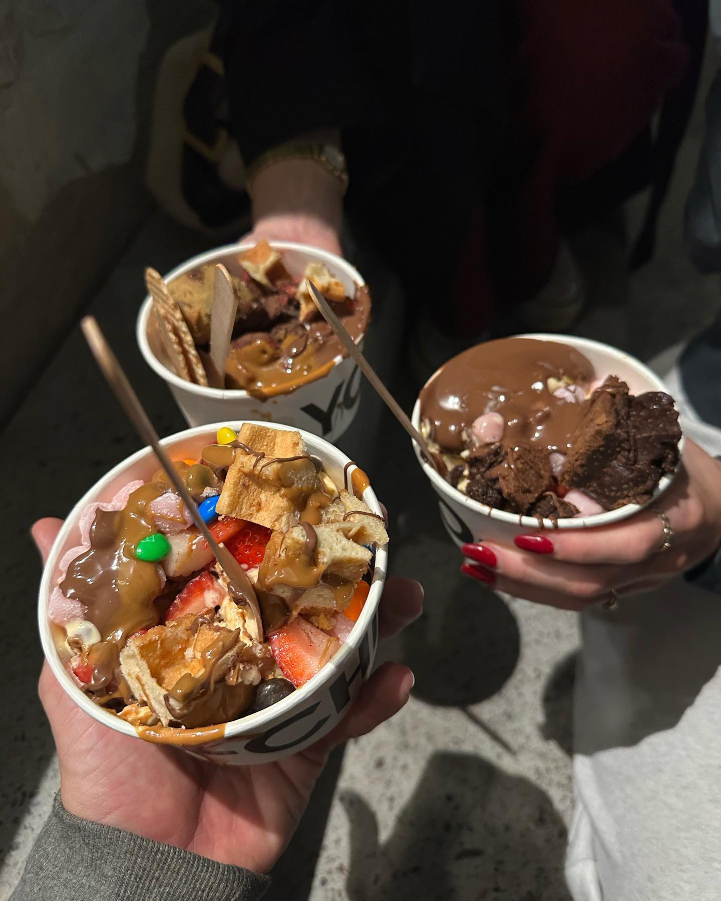 Three people are holding frozen yogurt cups with assorted toppings, including brownies, waffles, marshmallows, and strawberry pieces.