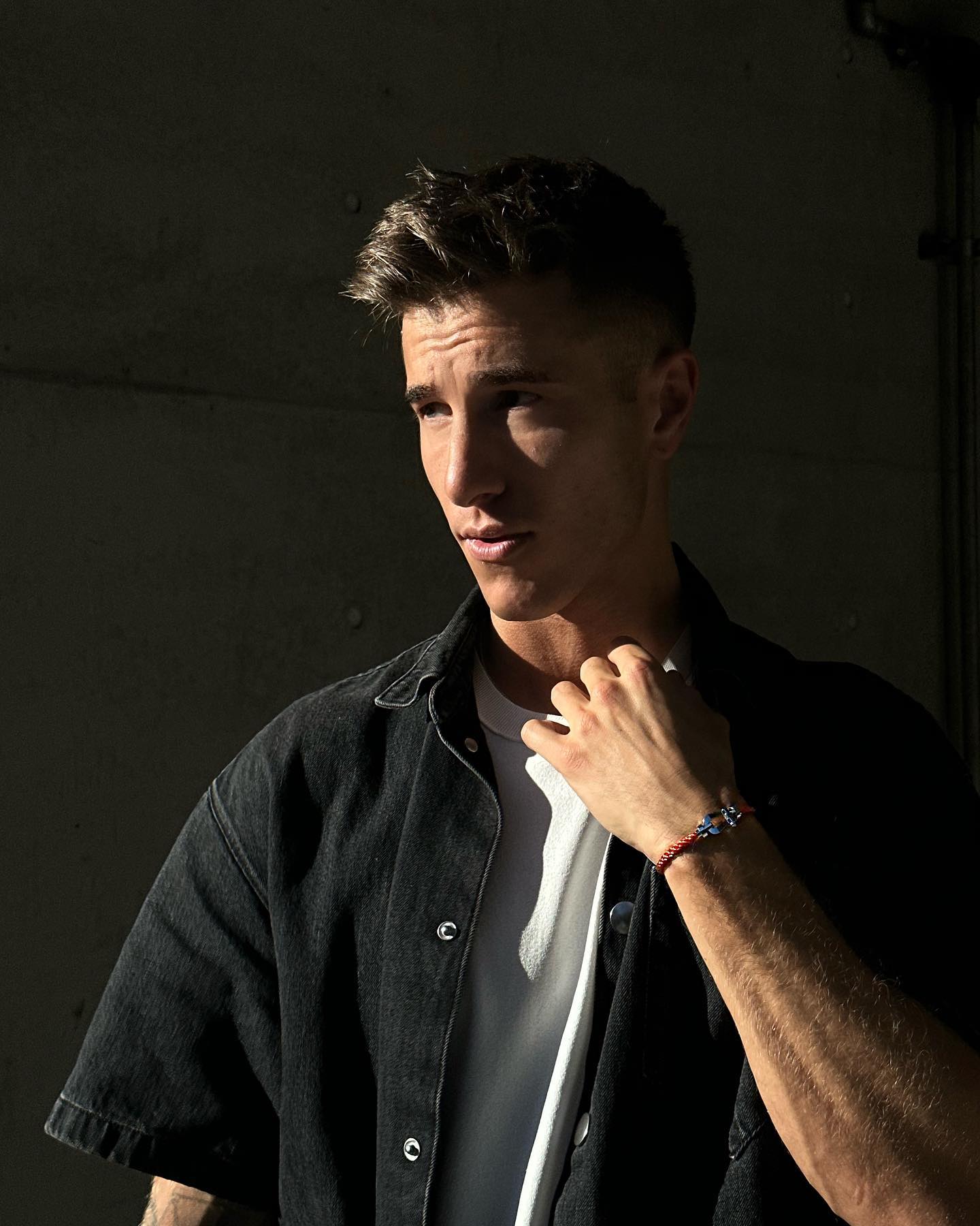 Portrait of a young adult male with short brown hair, wearing a black denim shirt and white t-shirt, posing in front of a dark wall.