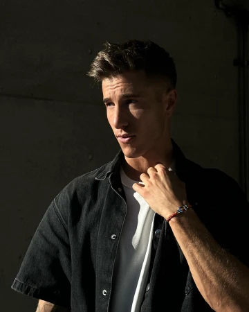 Portrait of a young adult male with short brown hair, wearing a black denim shirt and white t-shirt, posing in front of a dark wall.