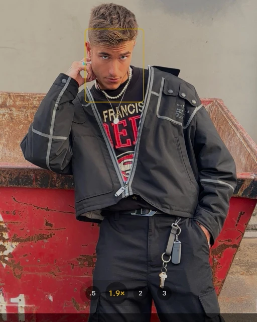 Stylish young man in black jacket and pants posing outdoors in front of rusty metal container with streetwear fashion details and accessories.