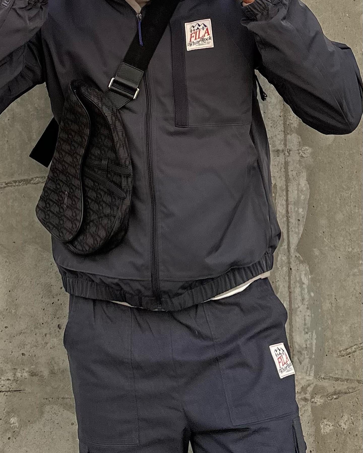 Person wearing a navy Fila tracksuit with a patterned Dior saddle bag in front of a gray concrete wall, fashion and street style.