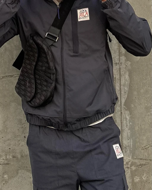 Person wearing a navy Fila tracksuit with a patterned Dior saddle bag in front of a gray concrete wall, fashion and street style.
