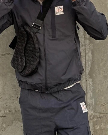 Person wearing a navy Fila tracksuit with a patterned Dior saddle bag in front of a gray concrete wall, fashion and street style.