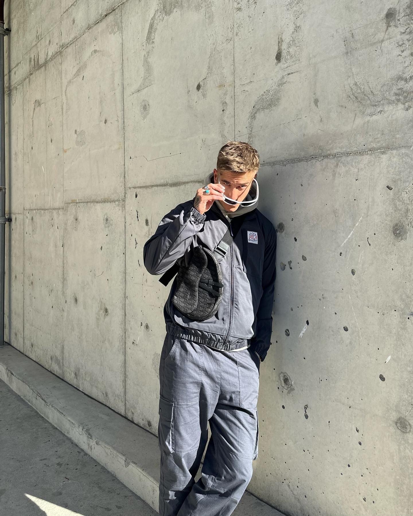 Blond male model posing against concrete wall in grey jacket and pants. He holds white-rimmed sunglasses with one hand in daytime.