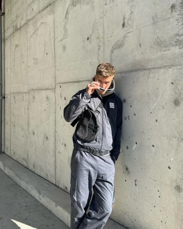 Man wearing grey track jacket and matching cargo pants leaning against a textured concrete wall, holding white sunglasses.