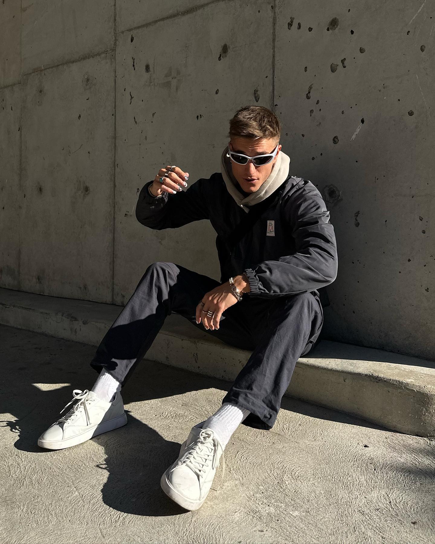 Adult male in sunglasses and black jacket sitting on a concrete step in an urban environment, wearing white sneakers and socks.