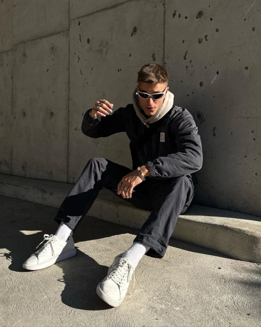 Adult male in sunglasses and black jacket sitting on a concrete step in an urban environment, wearing white sneakers and socks.