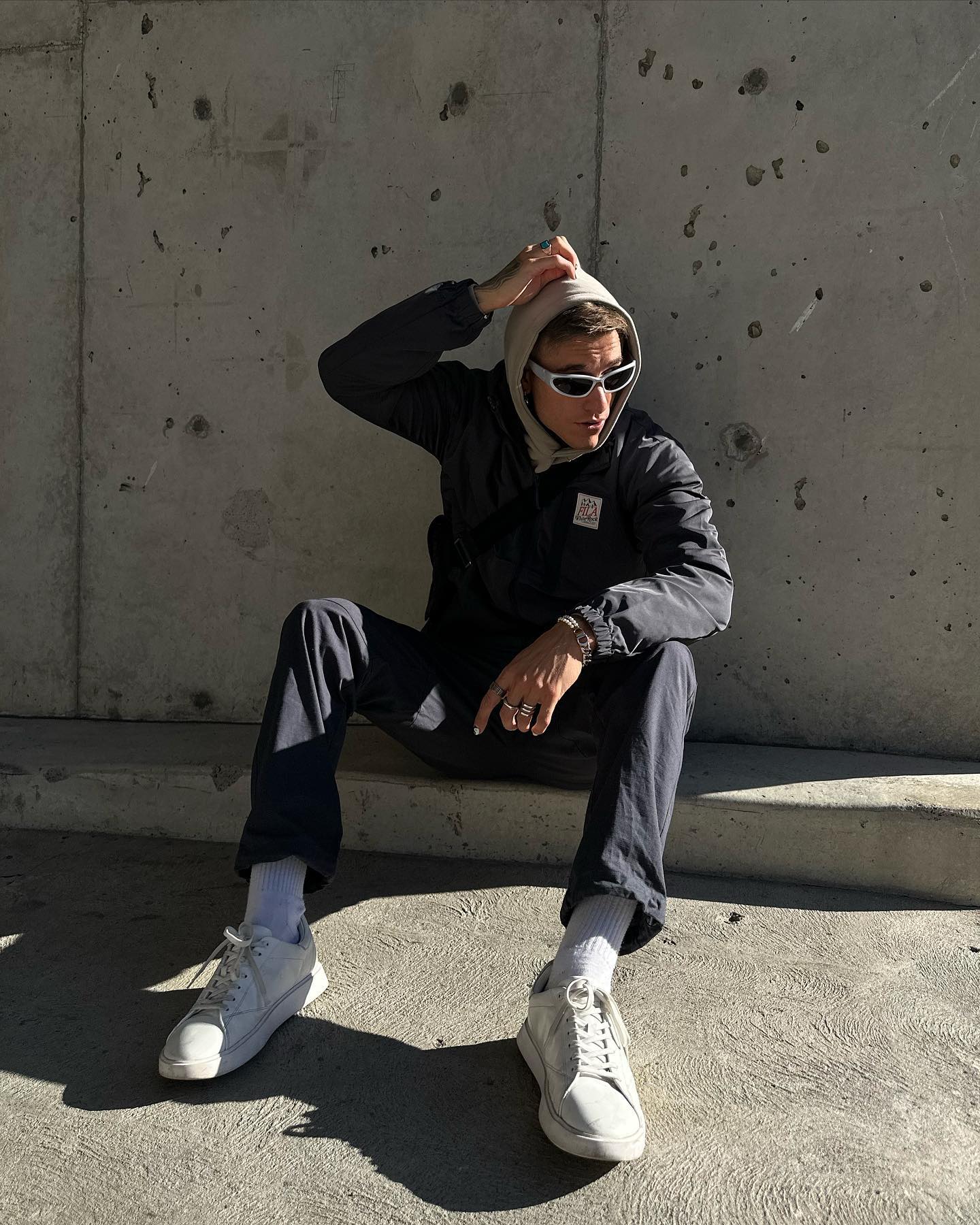 An adult male with sunglasses and a hooded sweatshirt sits in an urban setting, wearing casual attire and white sneakers, casting shadows.