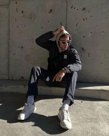 Man wearing sunglasses and a hoodie seated against a rough concrete wall outdoors in Sydney.