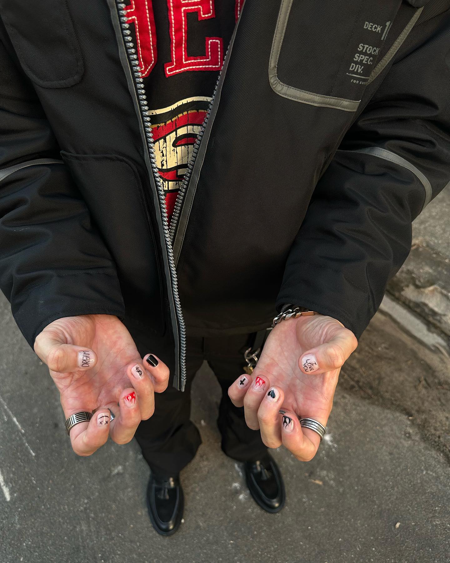 Close-up of a person's hands with nail art, rings, and a bracelet, also featuring their torso, jacket, shirt, pants, and shoes.
