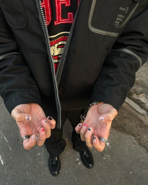 Close-up of a person's hands with nail art, rings, and a bracelet, also featuring their torso, jacket, shirt, pants, and shoes.