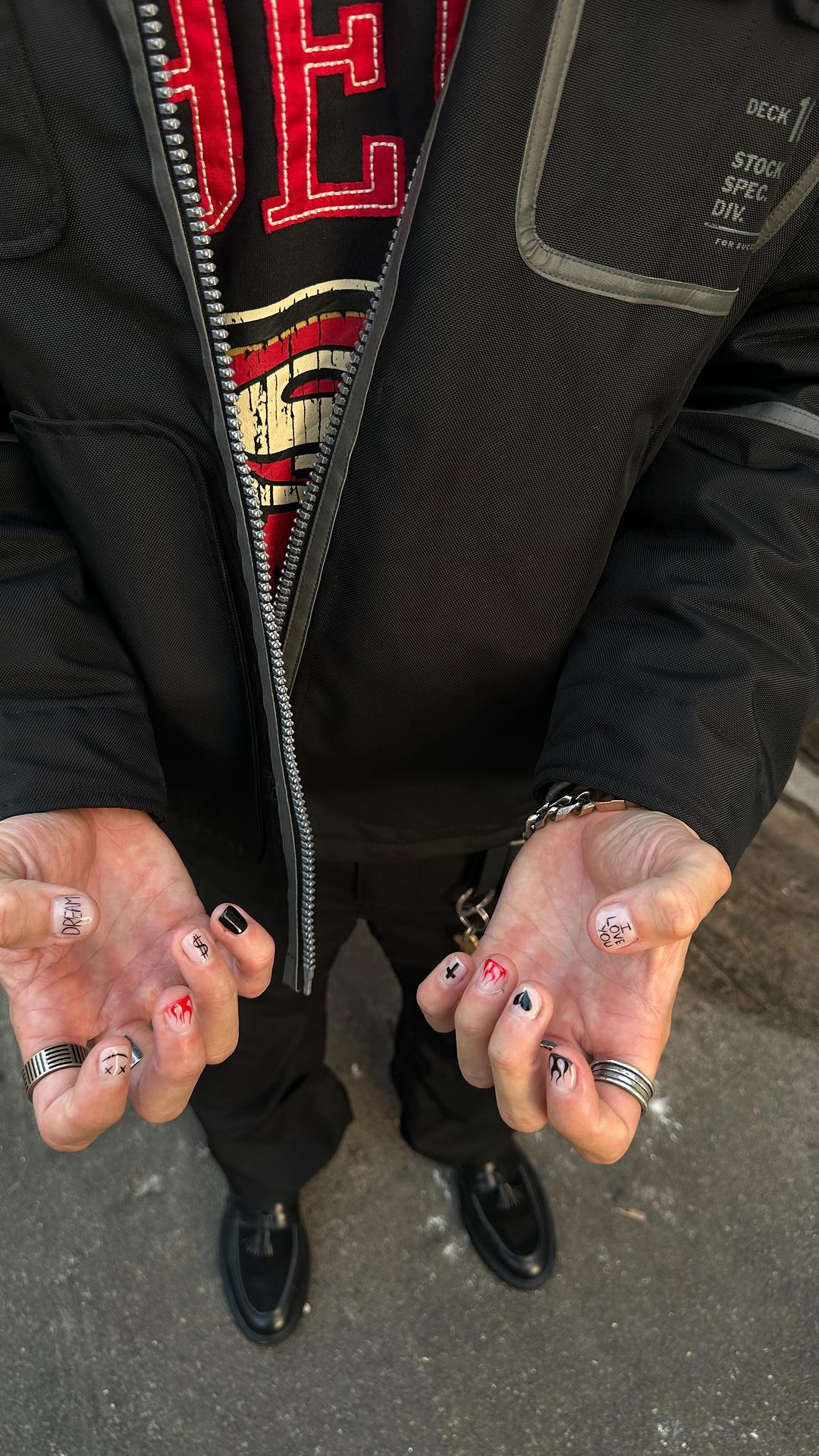 Man displays hands with nail art featuring black, red, and white designs. Rings adorn fingers, accessorizing casual street-style fashion.