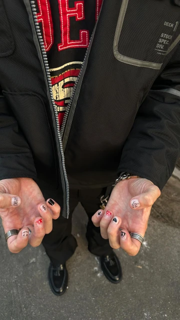 Man displays hands with nail art featuring black, red, and white designs. Rings adorn fingers, accessorizing casual street-style fashion.