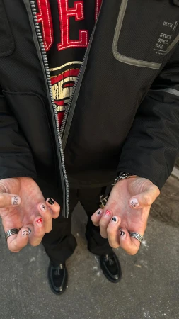 Man displays hands with nail art featuring black, red, and white designs. Rings adorn fingers, accessorizing casual street-style fashion.