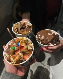 Three people holding paper cups filled with loaded ice cream desserts topped with chocolate sauce, fruit, and wafers.