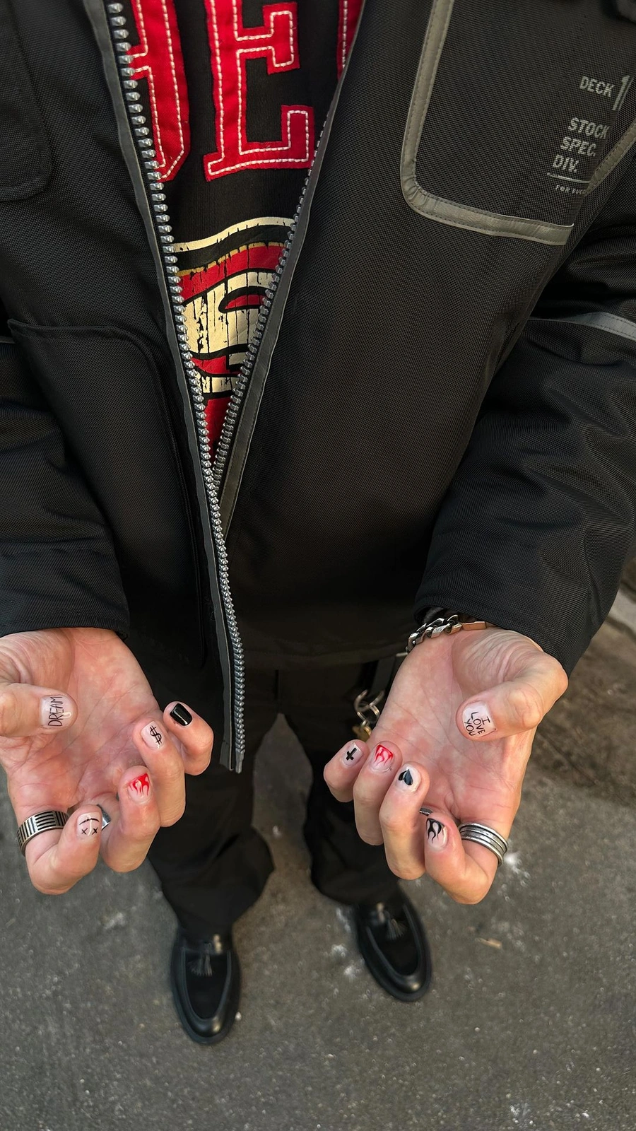 Close-up overhead shot of hands displaying custom nail art and rings, wearing a black jacket and graphic hoodie.