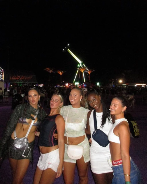 Five young women pose at an outdoor music festival at night, showcasing fashion and friendship in a group portrait with amusement park rides.