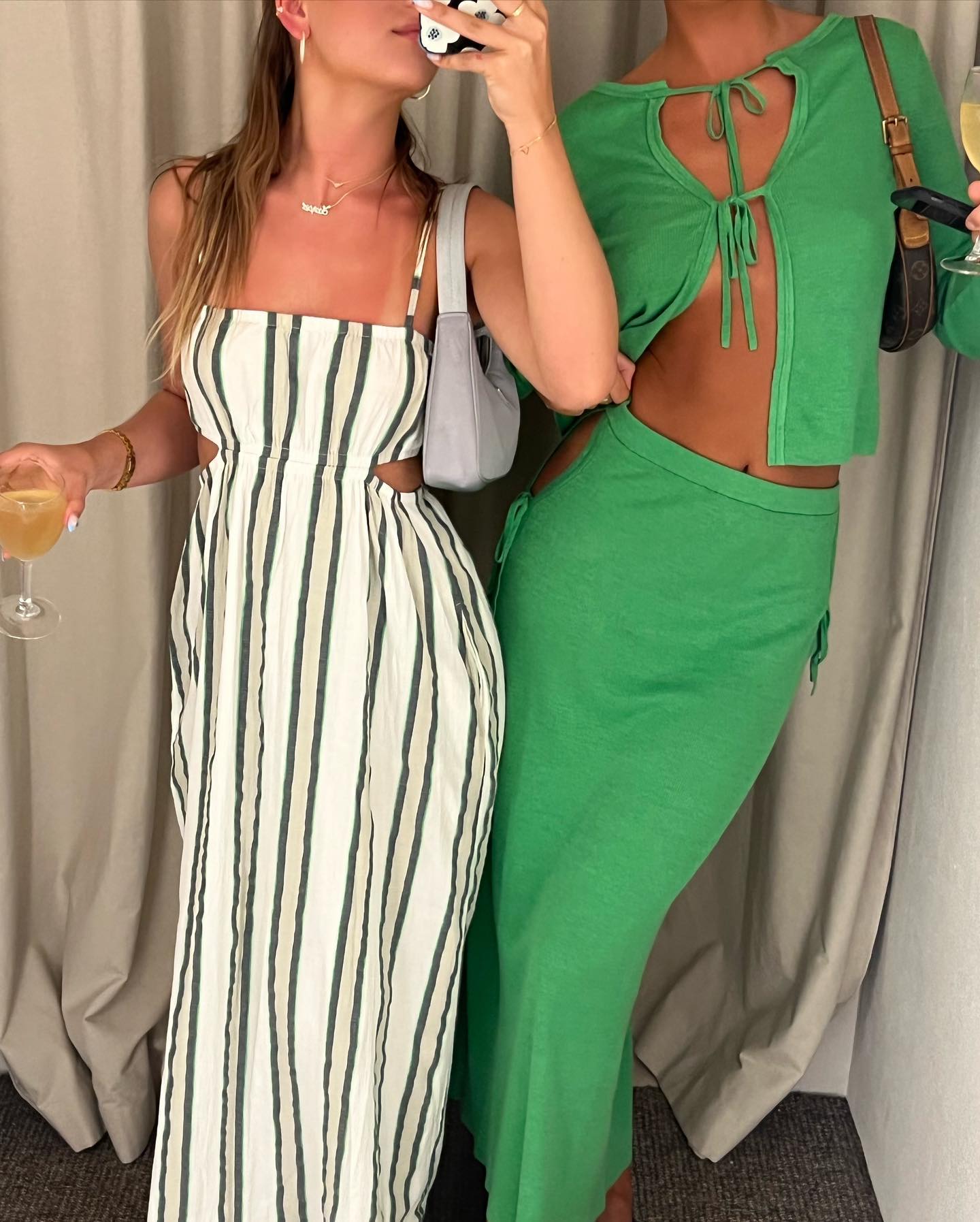 Two adult women in fashionable dresses pose indoors, one holding a wine glass and the other holding a smart phone. They are smiling.