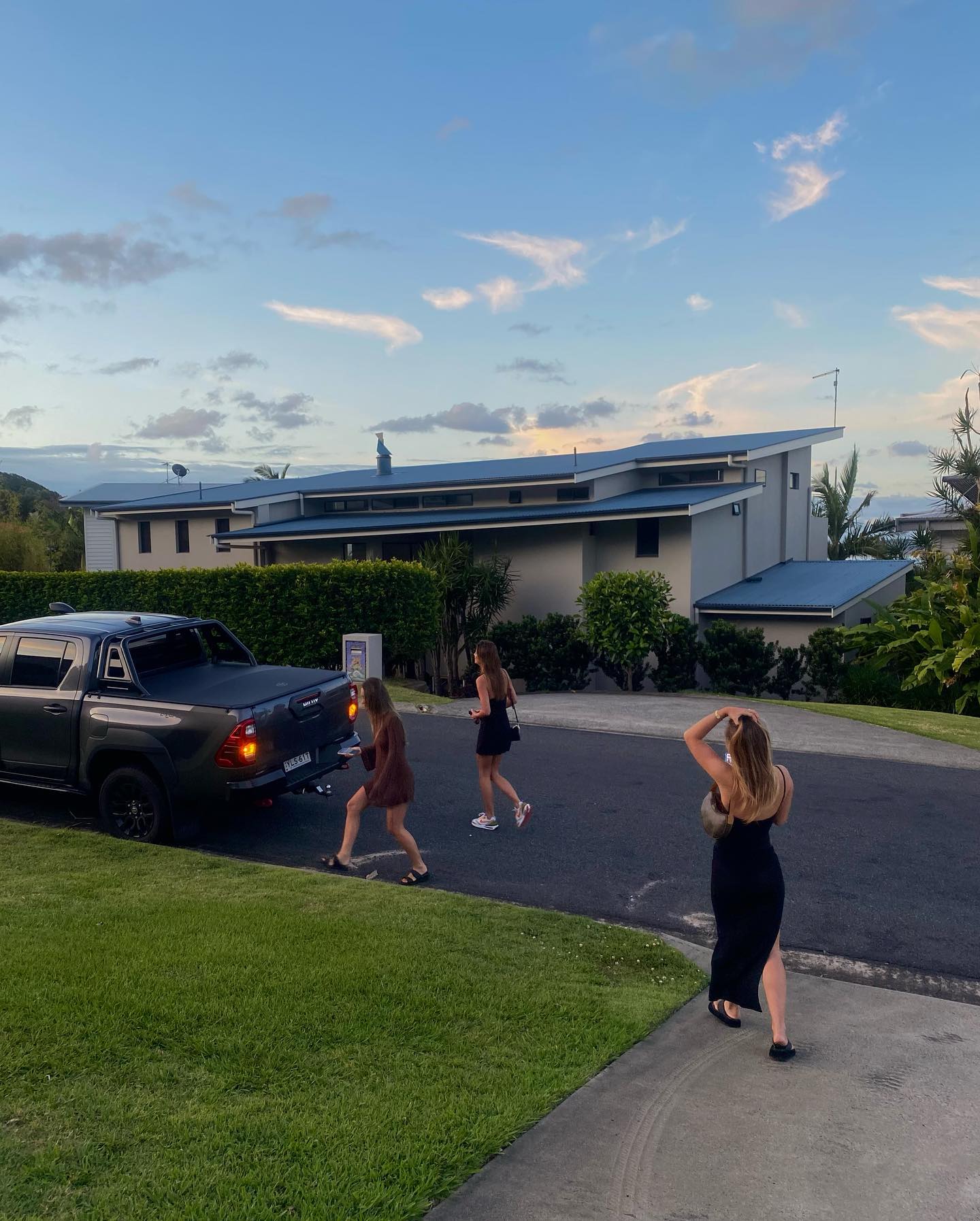 Three adult females walk away from a dark gray pick-up truck parked on an asphalt driveway in a suburban neighborhood at dusk.