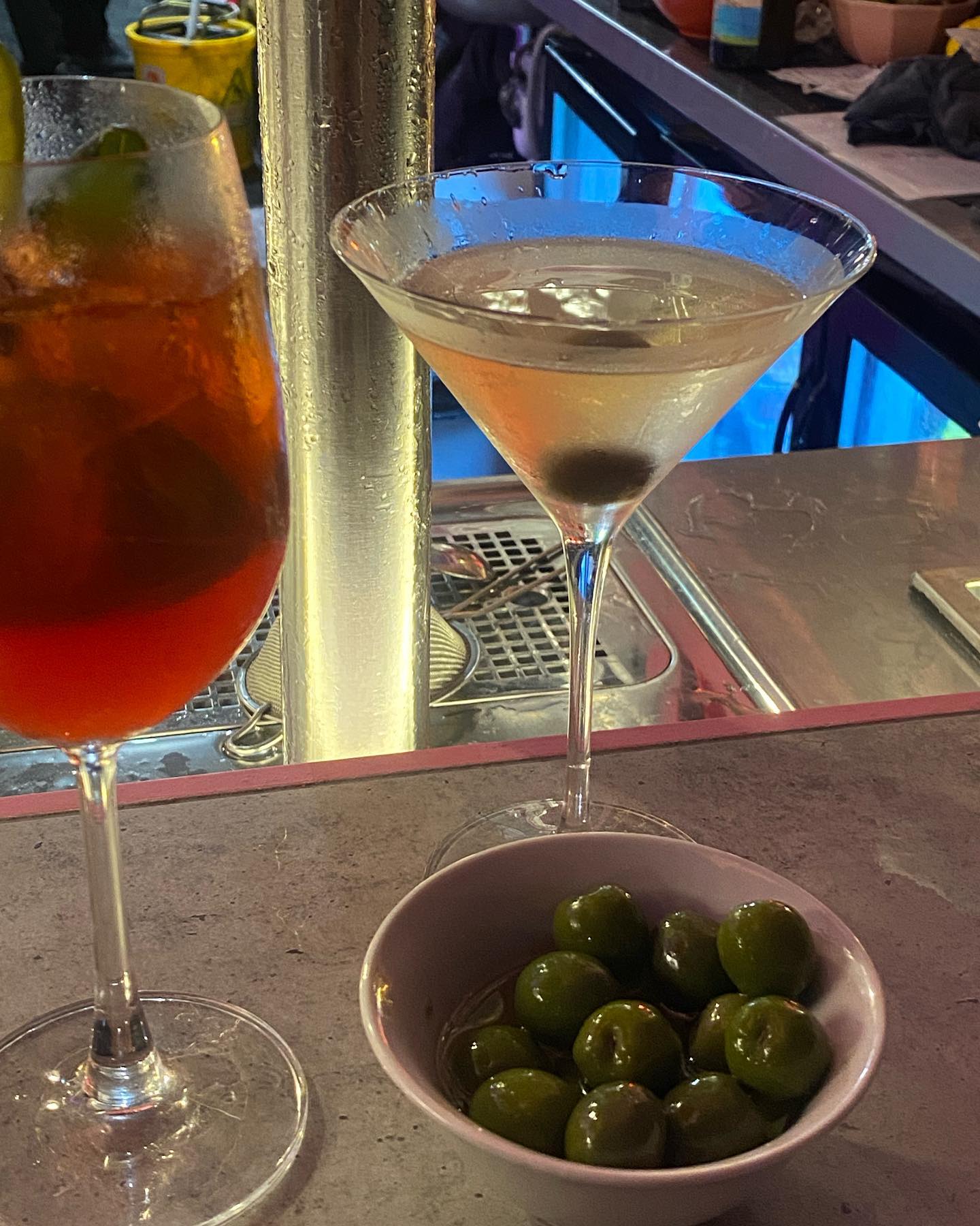 Two cocktails in stemmed glasses on a bar with a bowl of olives, featuring a martini and a red-colored beverage, illuminated by blue light.