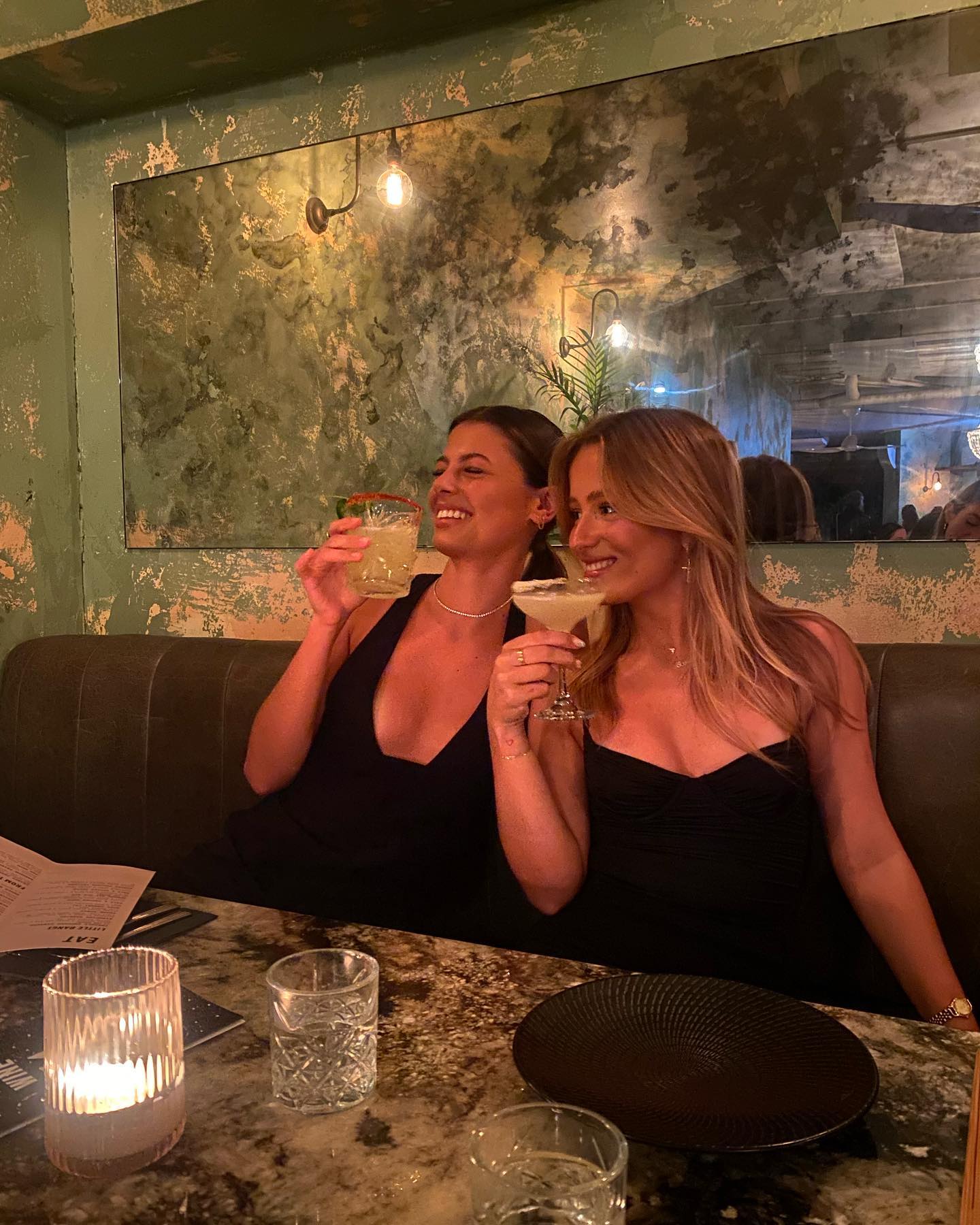 Two adult females smile and hold cocktails while sitting indoors at a restaurant, captured in a medium shot with warm lighting.