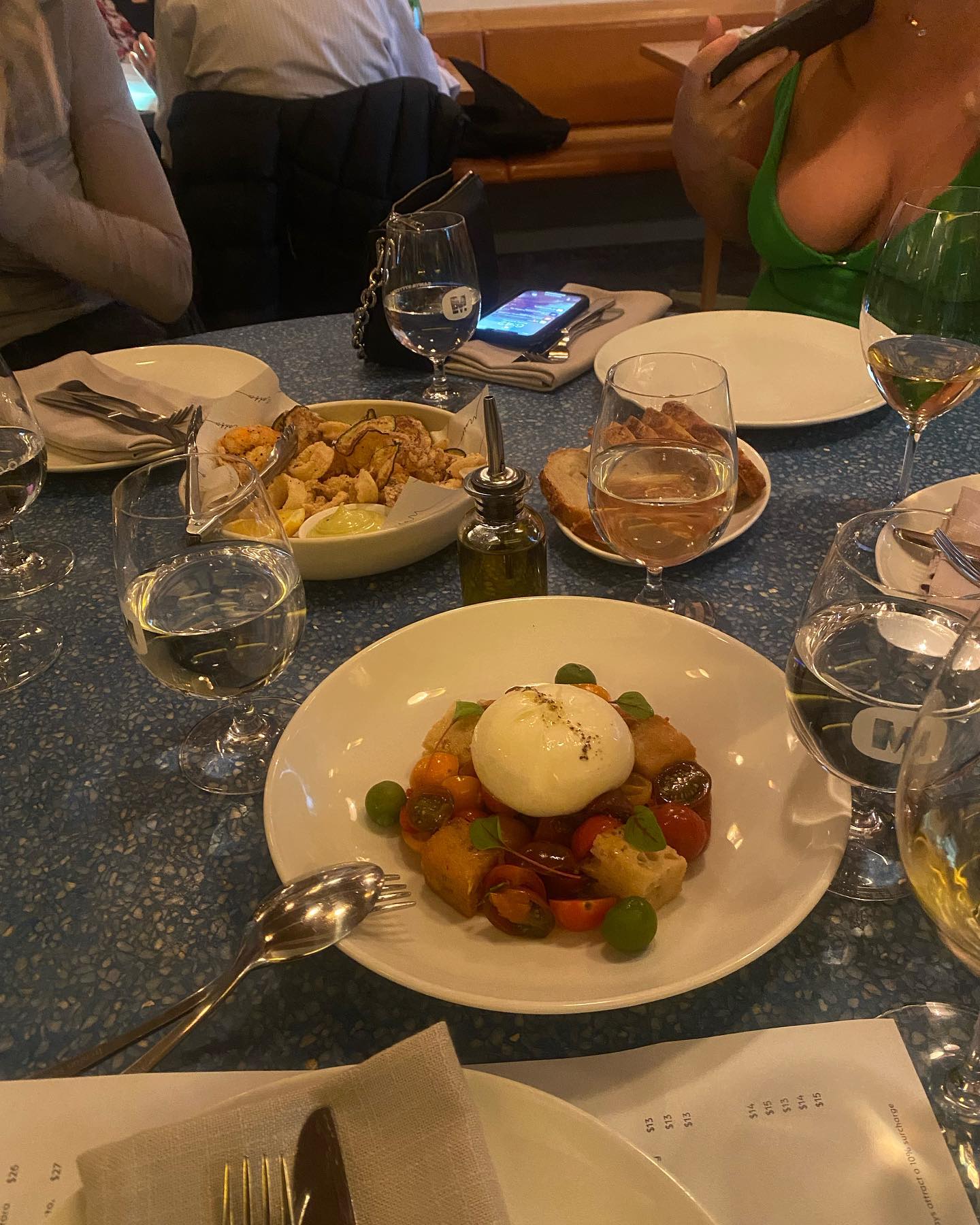 Table setting with burrata salad, fried seafood, and wine glasses in a restaurant with patrons in the background.